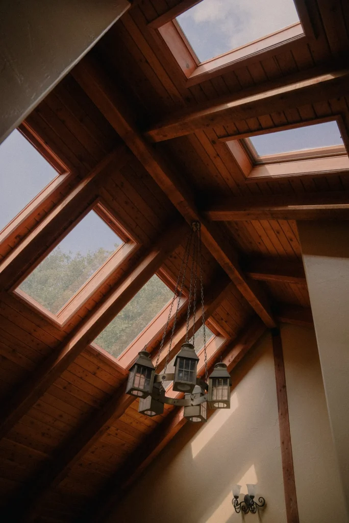 Wooden ceiling with multiple skylights, allowing soft daylight to illuminate the space. A vintage chandelier hangs prominently, adding rustic charm. How to Stop Skylight Condensation.