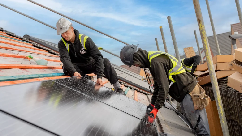 Two workers in safety gear install solar panels on a roof. They're focused and diligent under a clear blue sky, highlighting renewable energy efforts.