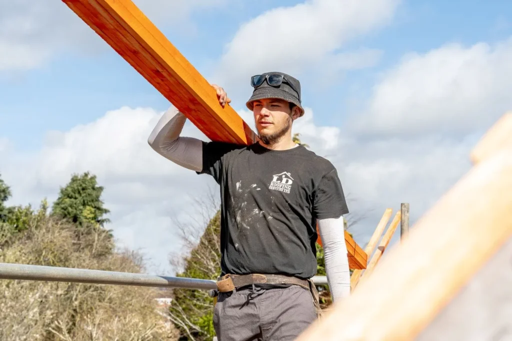A construction worker in a black shirt and bucket hat carries a wooden beam on his shoulder. Background shows trees and a cloudy blue sky. Confident expression.