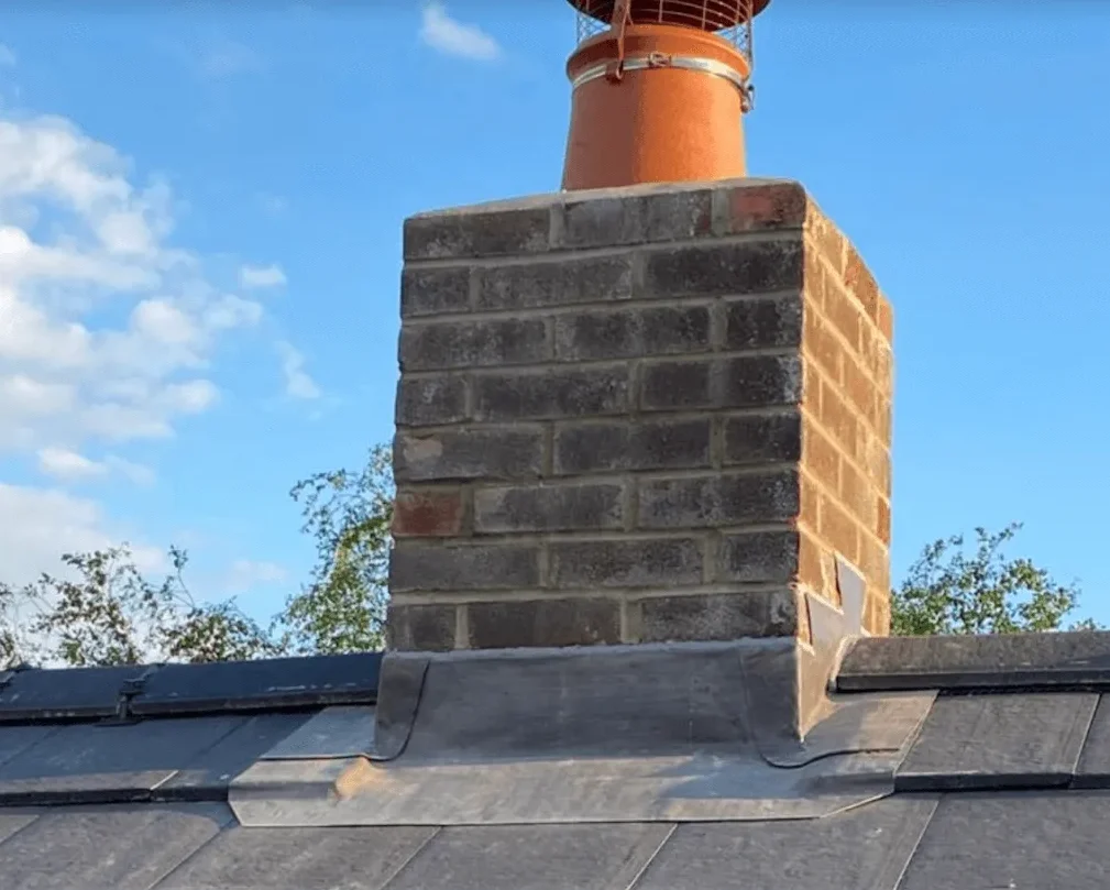 Brick chimney with metal flashing on a slate roof, under a clear blue sky. Trees are visible in the background. The scene conveys a serene, sunny day.