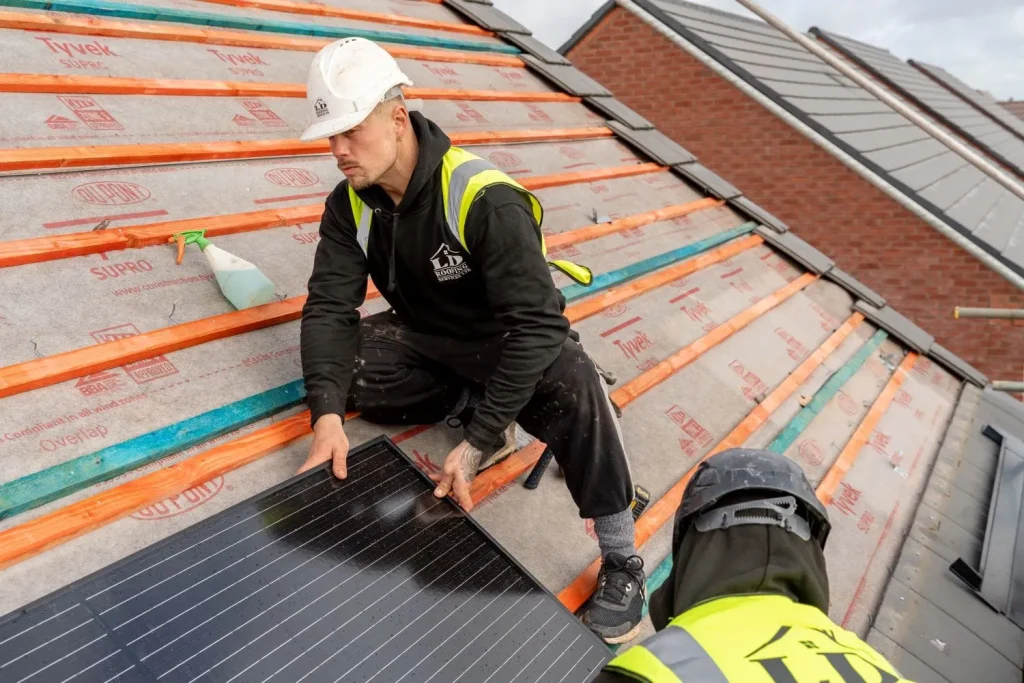 Two workers in safety gear install solar panels on a sloped roof. One aligns a panel, while the other assists, conveying teamwork and focus.