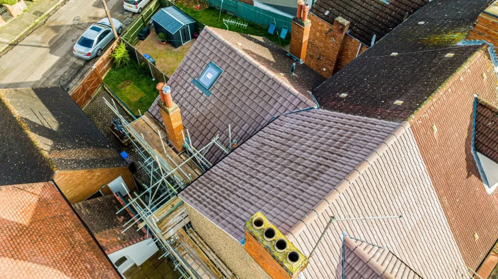 Aerial view of residential rooftops shows various brown shingles, a chimney, and a skylight. Scaffolding indicates roof maintenance in progress.